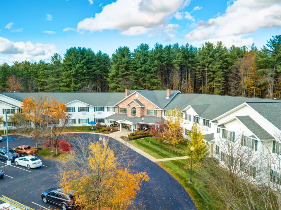 Aerial view of The Landing at Queensbury senior living facility showing a large two-story building with white siding and a brick entrance. The building is surrounded by trees with autumn foliage and a parking lot with several cars. The sky is partly cloudy with blue patches.