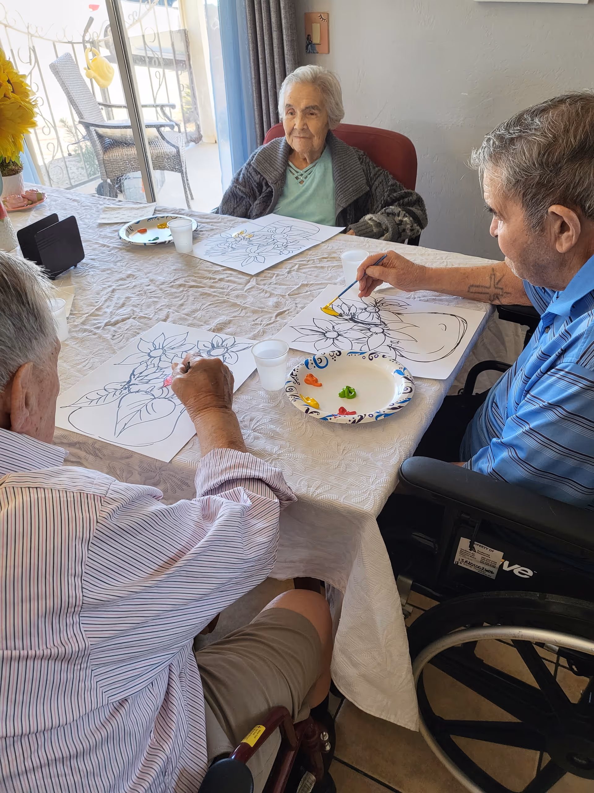 Three elderly individuals sitting around a table engaged in a painting activity with floral designs on paper. One person is in a wheelchair, and there are paint colors on a paper plate in the center of the table. The setting appears to be a bright room with a sliding glass door leading to an outdoor area.