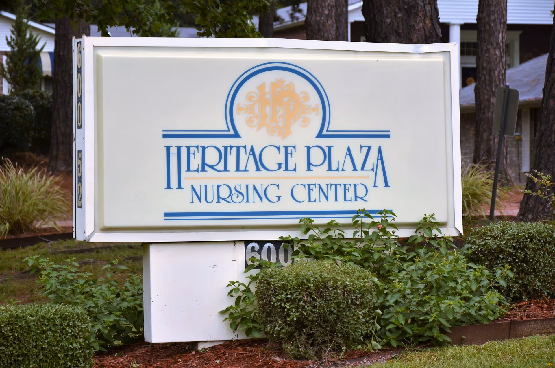 A freestanding sign that reads "Heritage Plaza Nursing Center" on a landscaped lawn with shrubs and trees behind it.