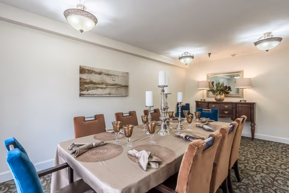 A formal dining room with a long table covered by a beige tablecloth, set with brown placemats, folded napkins, and gold-rimmed glasses. The table has silver candle holders with white candles. Surrounding the table are upholstered chairs in brown and blue. The room has beige walls, a patterned carpet, a sideboard with two lamps and a floral arrangement, and a large mirror above the sideboard.