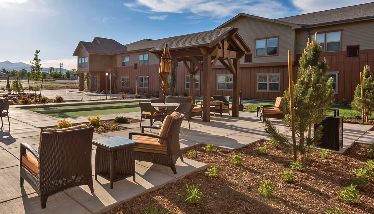 Outdoor patio area at Summit Senior Living featuring wicker chairs with orange cushions, small tables, a wooden pergola with a closed umbrella, landscaped garden beds with small plants and trees, and a two-story building in the background under a partly cloudy sky.