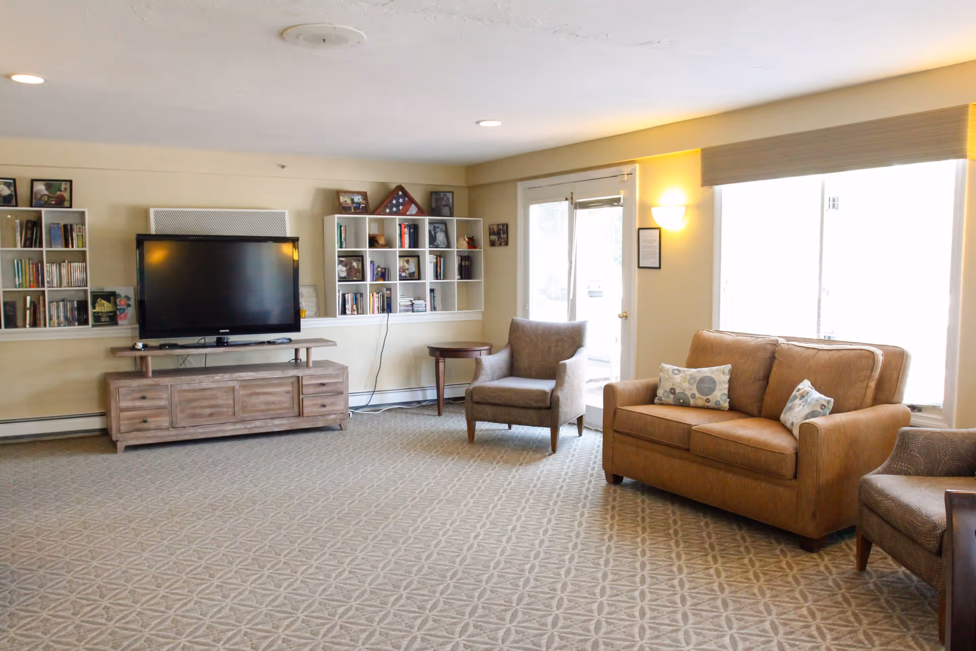 A bright living room with beige walls and carpeted floor featuring a brown leather loveseat with two patterned pillows, two gray armchairs, a wooden side table, and a wooden TV stand with a flat-screen television. Built-in white shelves with books and framed photos are mounted on the wall behind the TV. Large windows and a glass door allow natural light to fill the room.