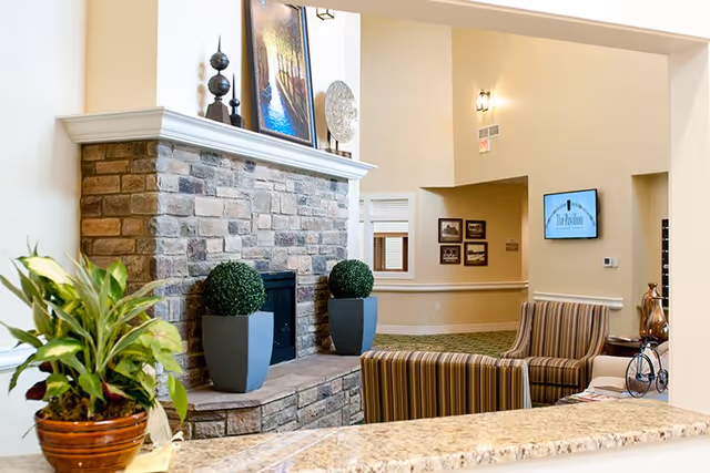 A cozy interior common area featuring a stone fireplace with decorative plants on the hearth and artwork on the mantel. In front of the fireplace are two striped armchairs and a small table with a decorative bicycle sculpture. A potted plant is visible on a granite countertop in the foreground. The wall in the background has framed pictures and a mounted TV screen displaying The Pavilion Senior Living logo.