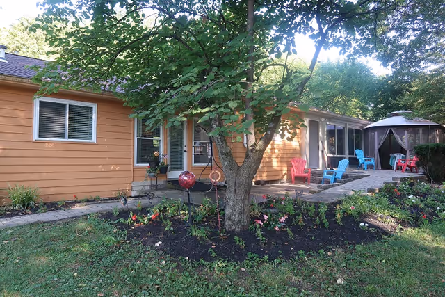 One-story orange house exterior with a tree, flowerbed and a stone patio furnished with colorful Adirondack chairs and a gazebo.