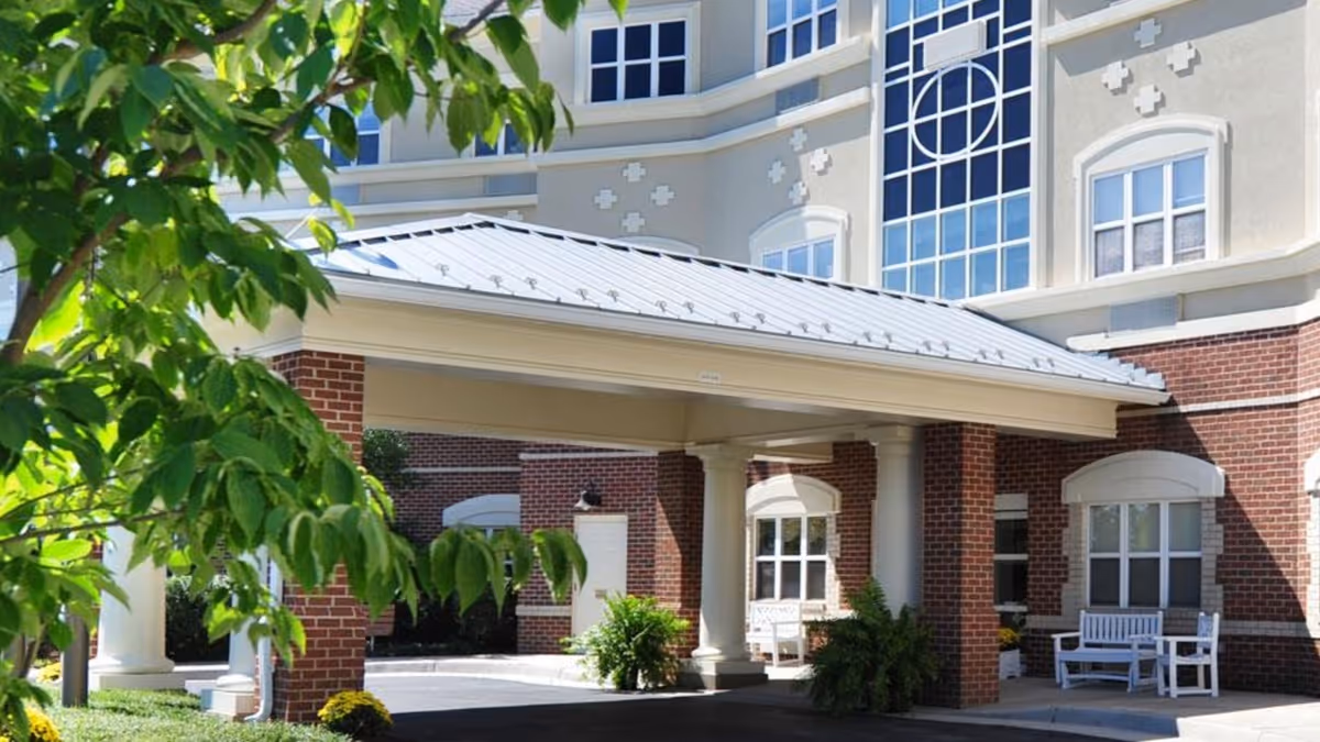 Entrance area of a senior living facility with a covered drop-off zone supported by white columns and brick walls. There are white benches and green plants near the entrance, with a tree partially visible in the foreground. The building has multiple windows and decorative architectural details.