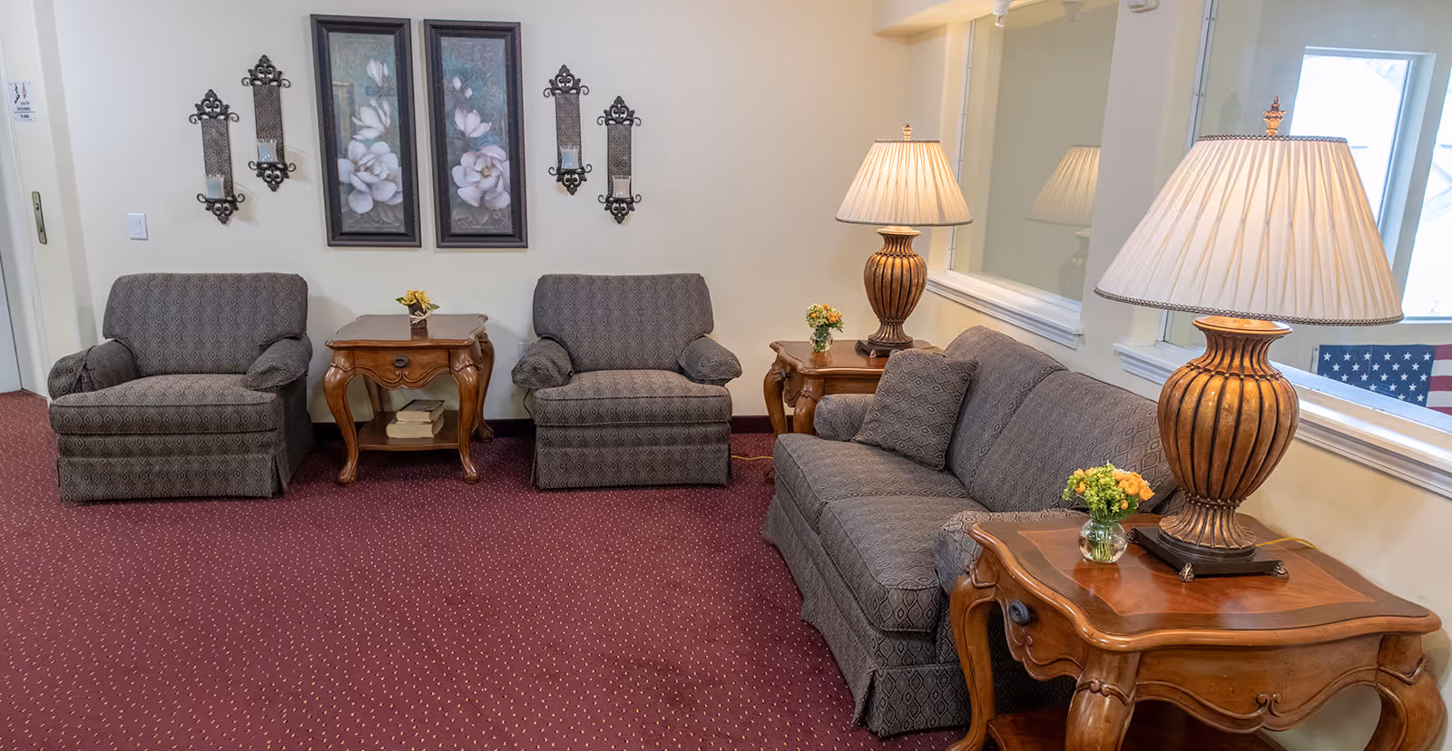 A cozy sitting area with two armchairs and a sofa upholstered in patterned gray fabric. There are two wooden side tables with decorative lamps and small flower arrangements. The walls are adorned with floral artwork and candle sconces. The floor is covered with a red carpet with small yellow dots.