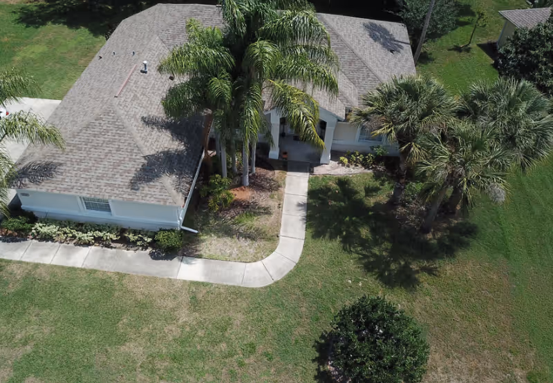 Aerial view of a single-story building with a brown shingled roof surrounded by green grass and palm trees. A concrete walkway leads to the entrance of the building, which is partially shaded by the trees.