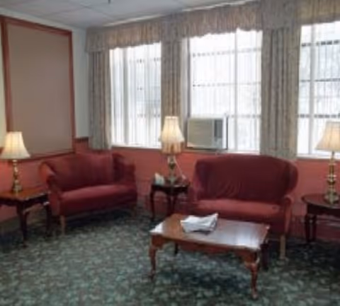 A cozy sitting area with two red upholstered loveseats, a wooden coffee table with a folded cloth on it, two side tables each with a lamp, and large windows with curtains and an air conditioning unit.