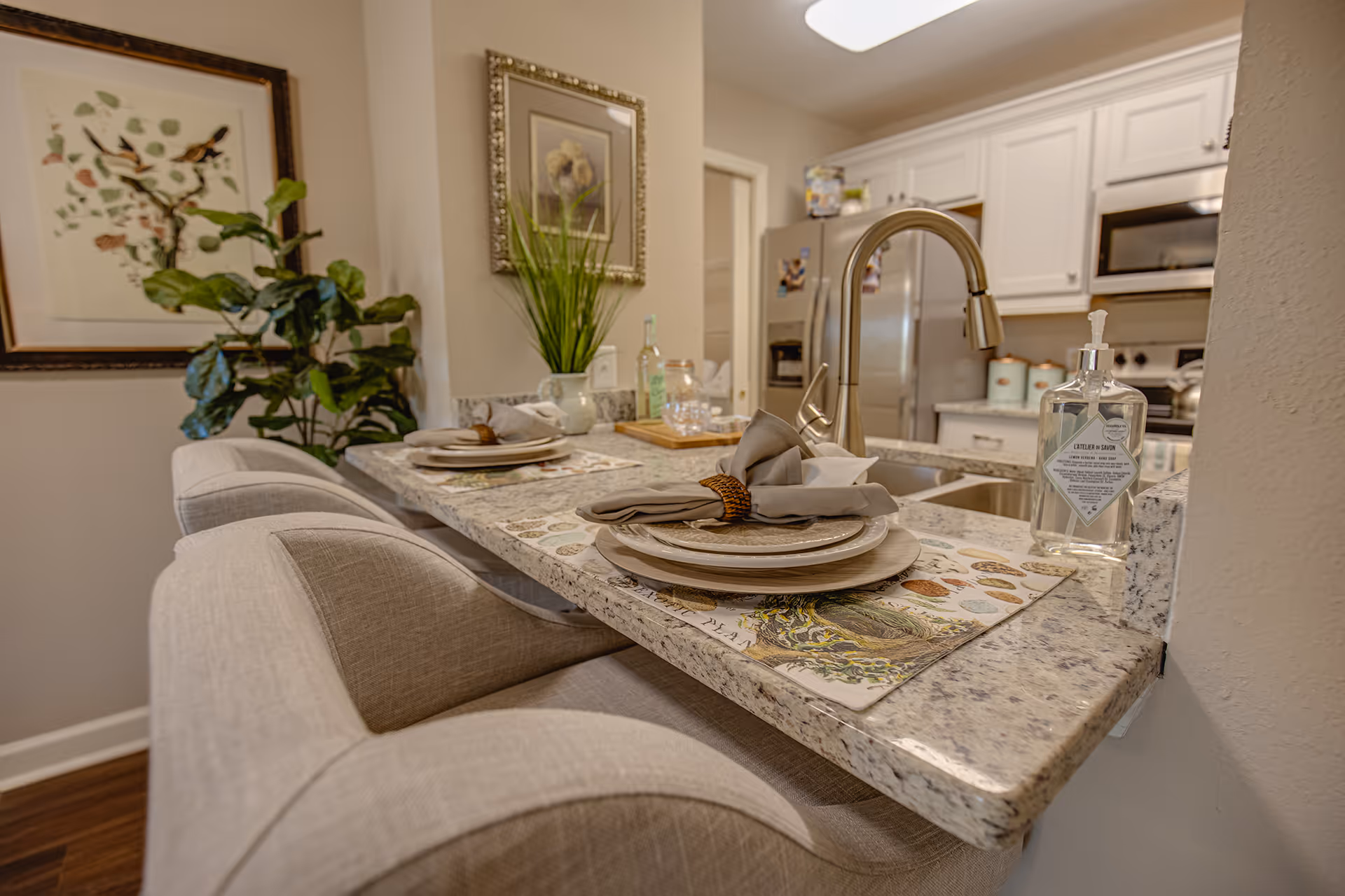 A granite kitchen breakfast bar with place settings, upholstered bar stools, a sink faucet, and white cabinets and refrigerator in the background.