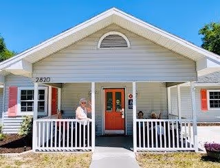 Front exterior view of a single-story assisted living facility named Coral Landing Assisted Living, featuring a covered porch with white railings, a red front door, and a person standing on the porch. The building is light-colored with red shutters and the address number 2820 is visible near the entrance.