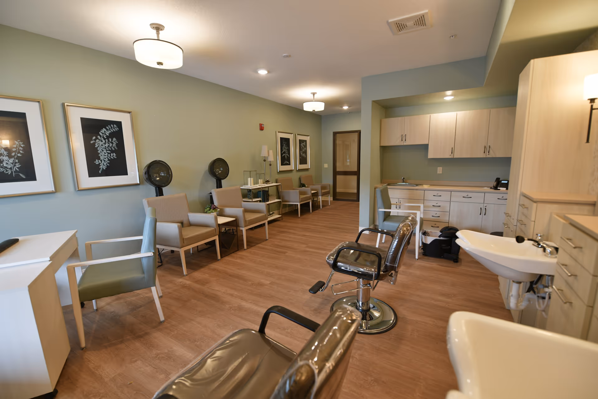 Interior view of a salon area in a senior living facility with salon chairs covered in plastic, hair drying stations, beige armchairs along the wall, framed botanical prints, and light wood cabinetry with a sink.
