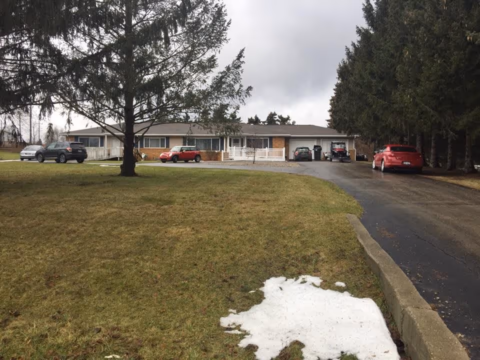 Exterior view of a single-story retirement facility building with a driveway and several parked cars. There is a large grassy area with a patch of snow in the foreground and tall trees on the right side. The sky is overcast.