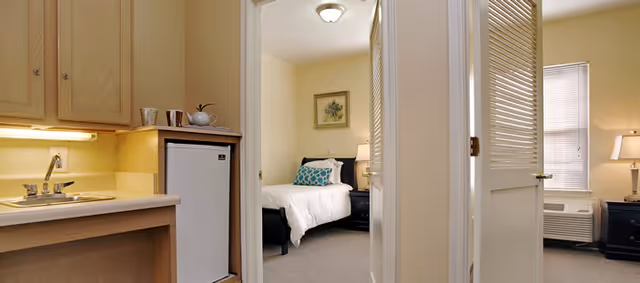 Interior view showing a small kitchenette on the left and a bedroom through an open door with a bed, nightstand, and window.