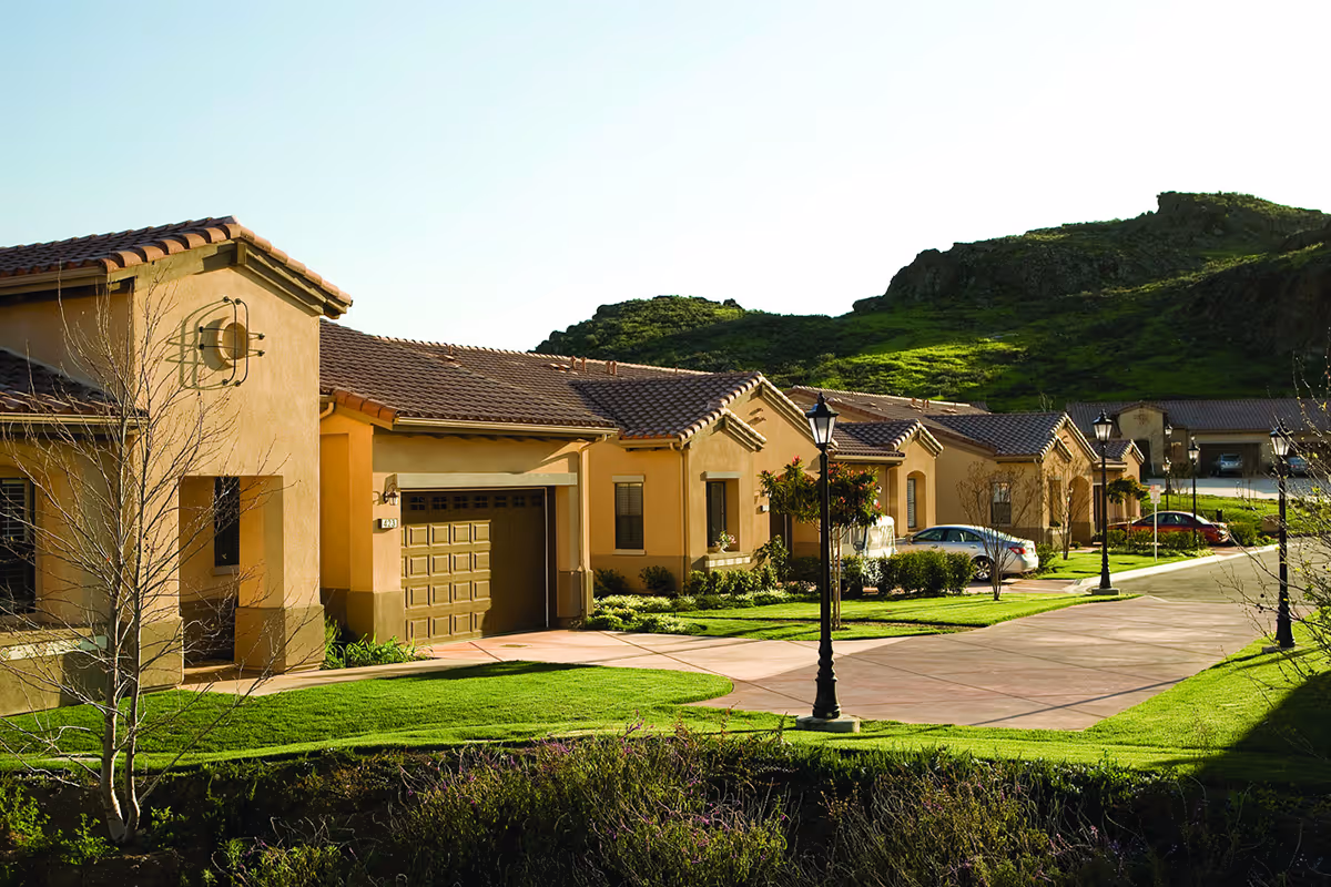 Row of single-story residential buildings with tan stucco walls and red tile roofs, set along a paved driveway with street lamps and green lawns, with hills in the background under a clear sky.
