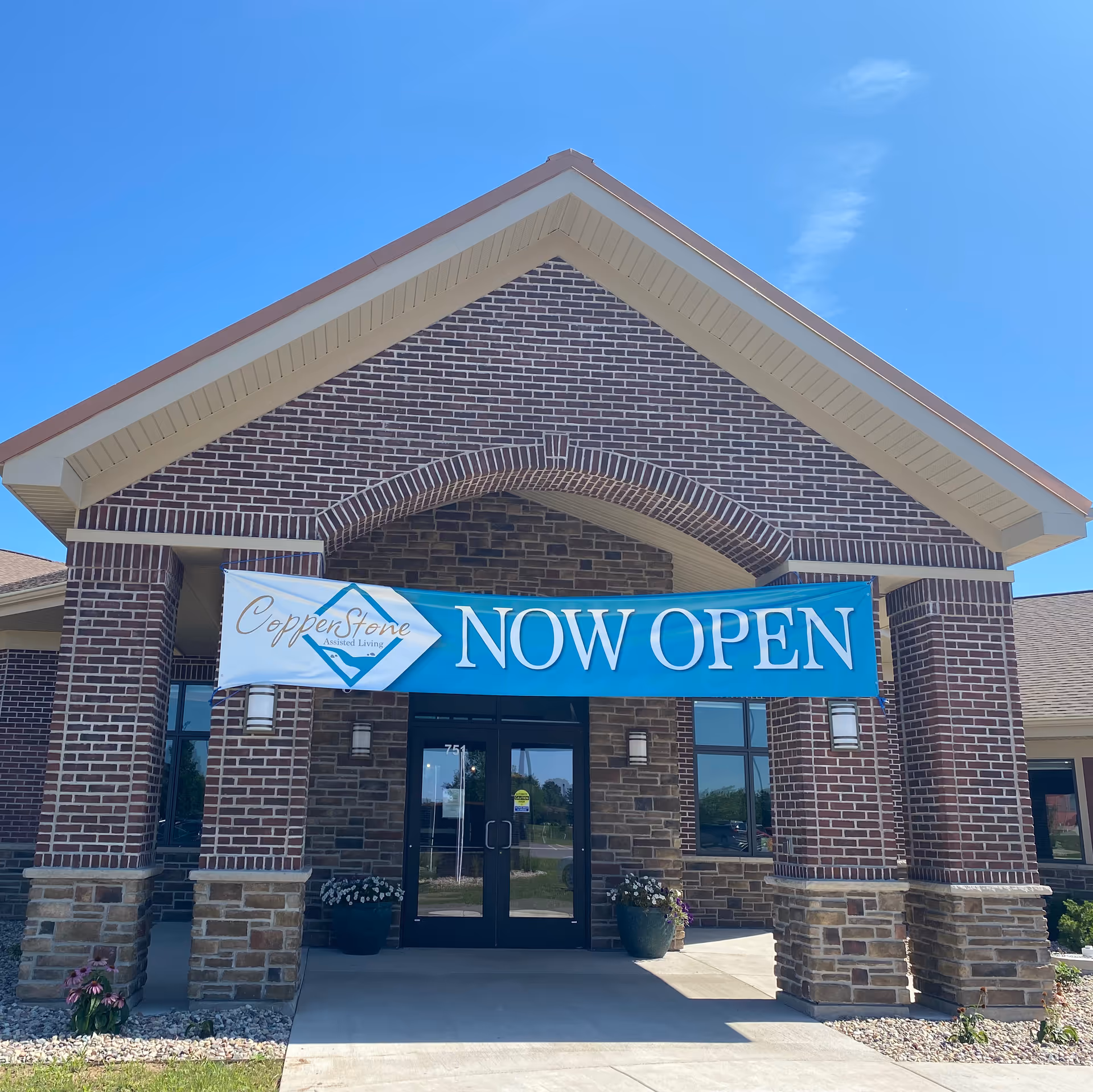 Front entrance of a brick building with a large blue banner that reads 'CopperStone Assisted Living NOW OPEN' hanging above the double glass doors. There are two large planters with flowers on either side of the entrance and a clear blue sky in the background.