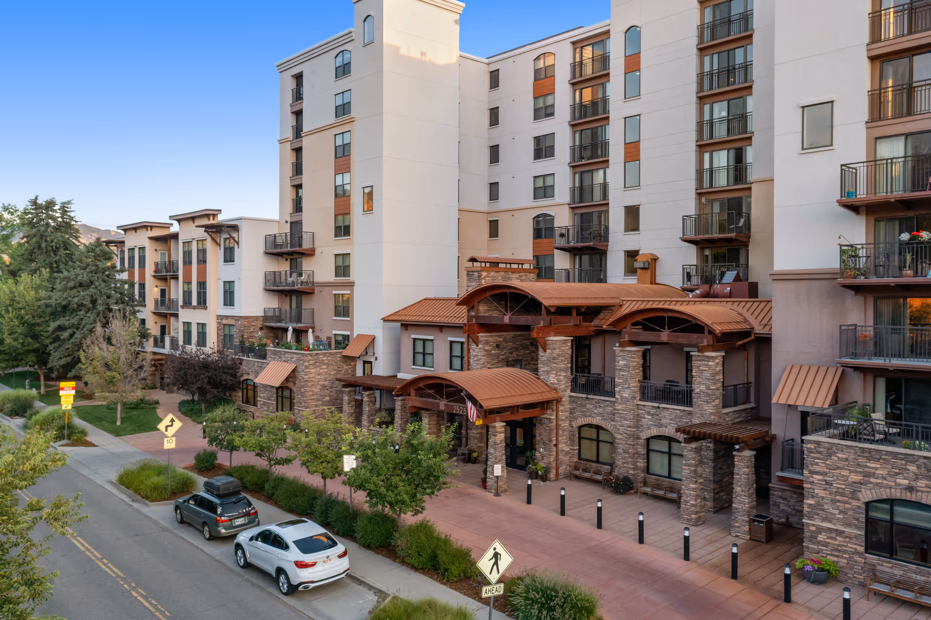 Exterior view of a multi-story senior living facility with balconies, stone and stucco facade, and a covered entrance. There are trees, shrubs, and parked cars along the street in front of the building under a clear blue sky.