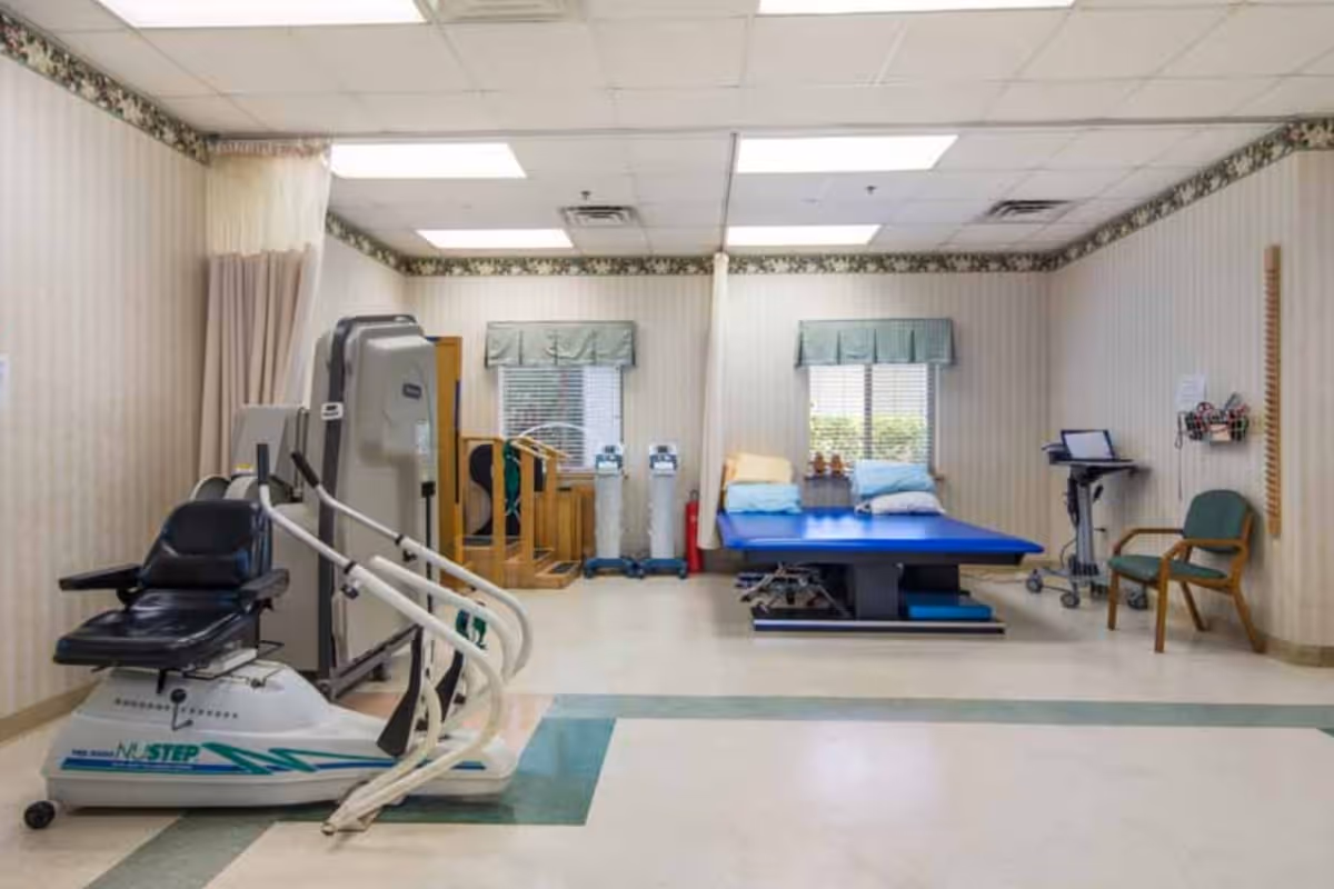 A physical therapy or rehabilitation room with exercise equipment including a seated stepper machine, parallel bars for walking practice, a large blue therapy table with pillows, two chairs, and medical equipment. The room has beige striped wallpaper with floral border near the ceiling, two windows with green valances, and fluorescent ceiling lights.