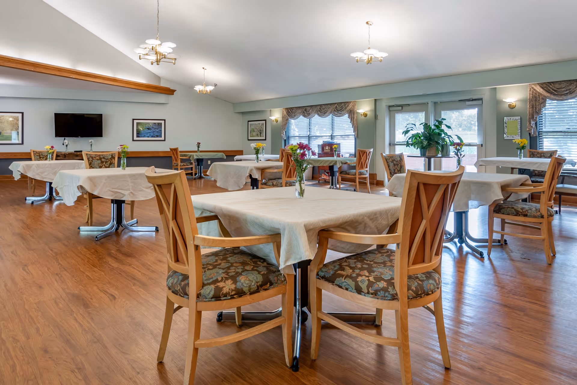 Bright communal dining room with multiple tables covered in cloths, wooden chairs with patterned cushions, small floral centerpieces, and large windows.