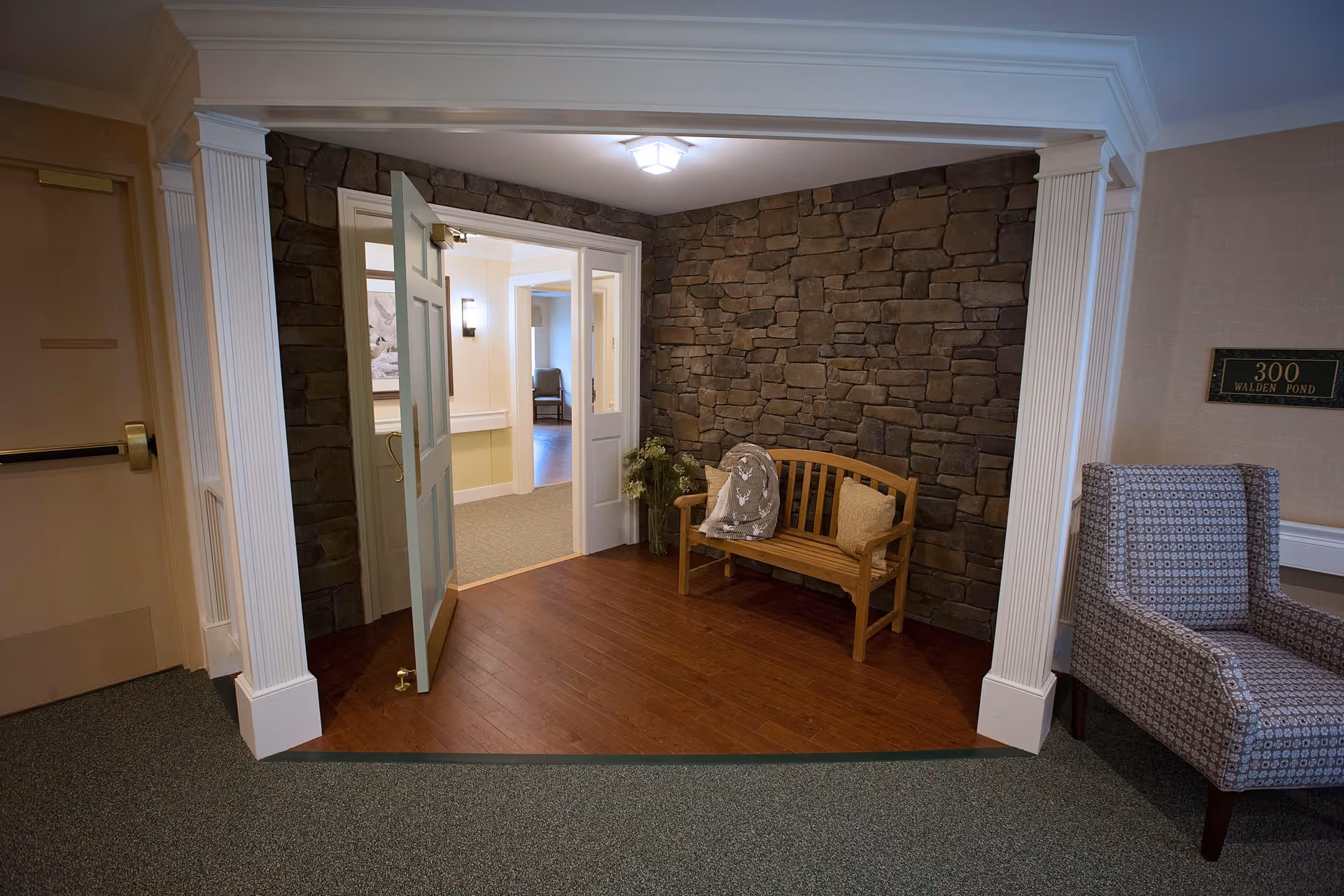 A cozy seating nook in a senior living facility featuring a wooden bench with a cushion and a decorative pillow, a patterned armchair, and a stone accent wall. The area has wooden flooring and is framed by white columns. An open door leads to a well-lit hallway with additional seating visible.
