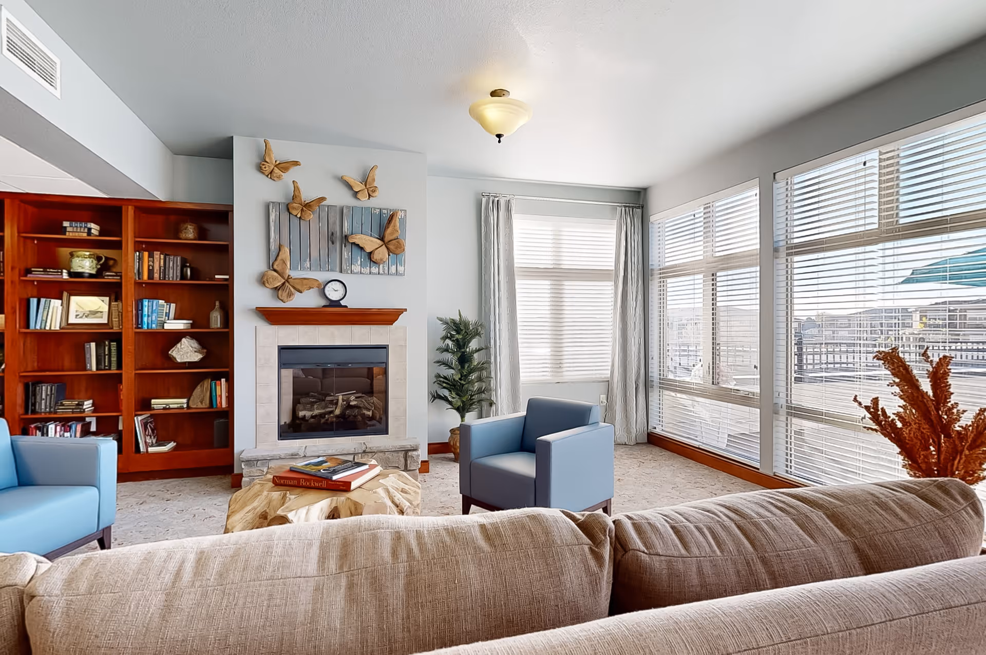 Bright living room with a fireplace, built-in bookshelf, sofa and blue armchairs by large windows with blinds.