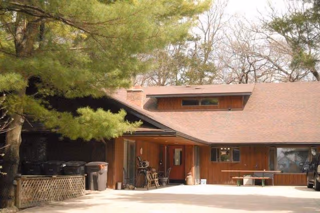 Exterior view of a single-story building with a sloped roof surrounded by trees. The building has wooden siding and a covered entrance area with chairs and a table. There are trash bins on the left side and a bench on the right side near a window.