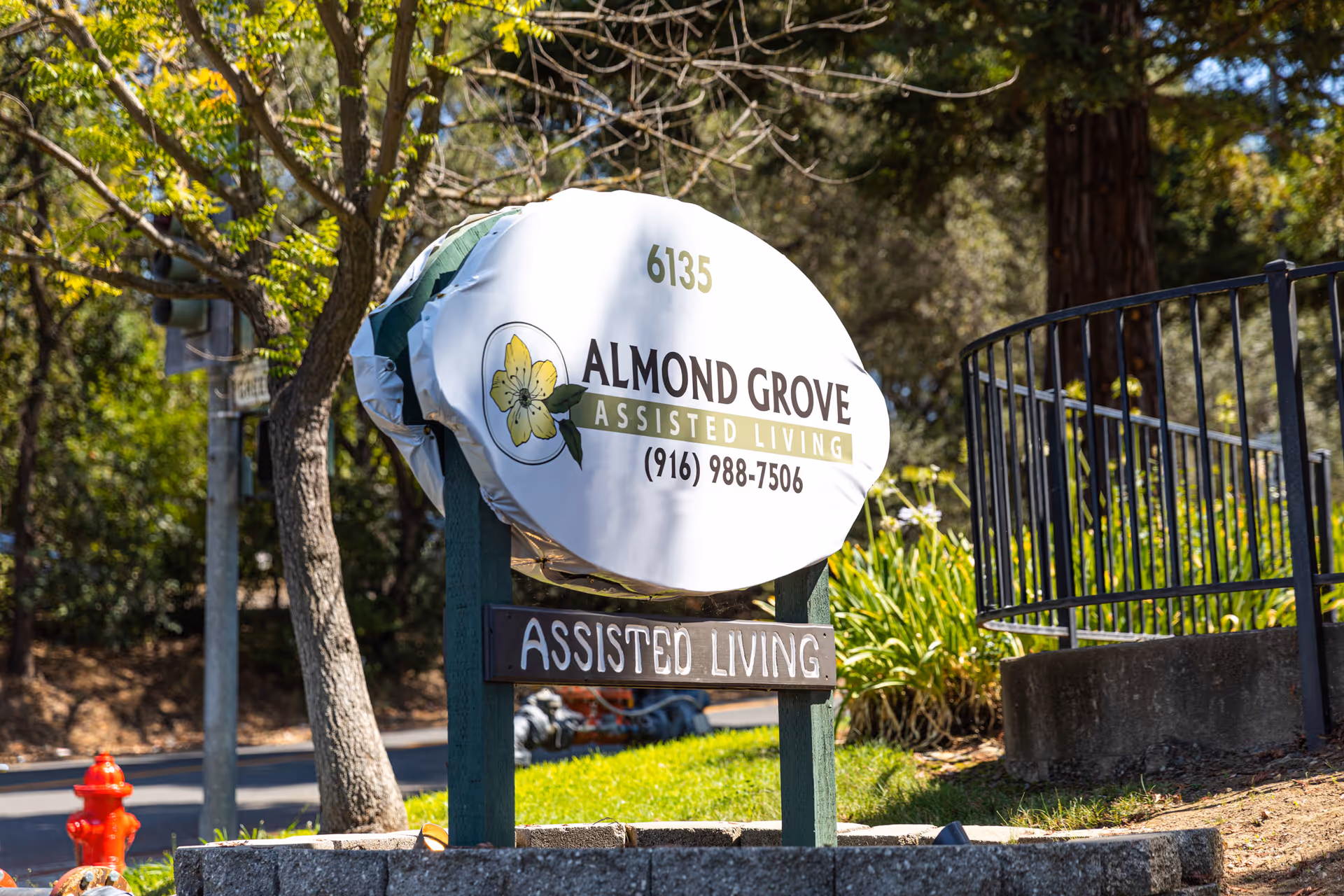 Outdoor sign for Almond Grove Assisted Living with the address number 6135 and phone number (916) 988-7506, surrounded by trees and greenery on a sunny day.