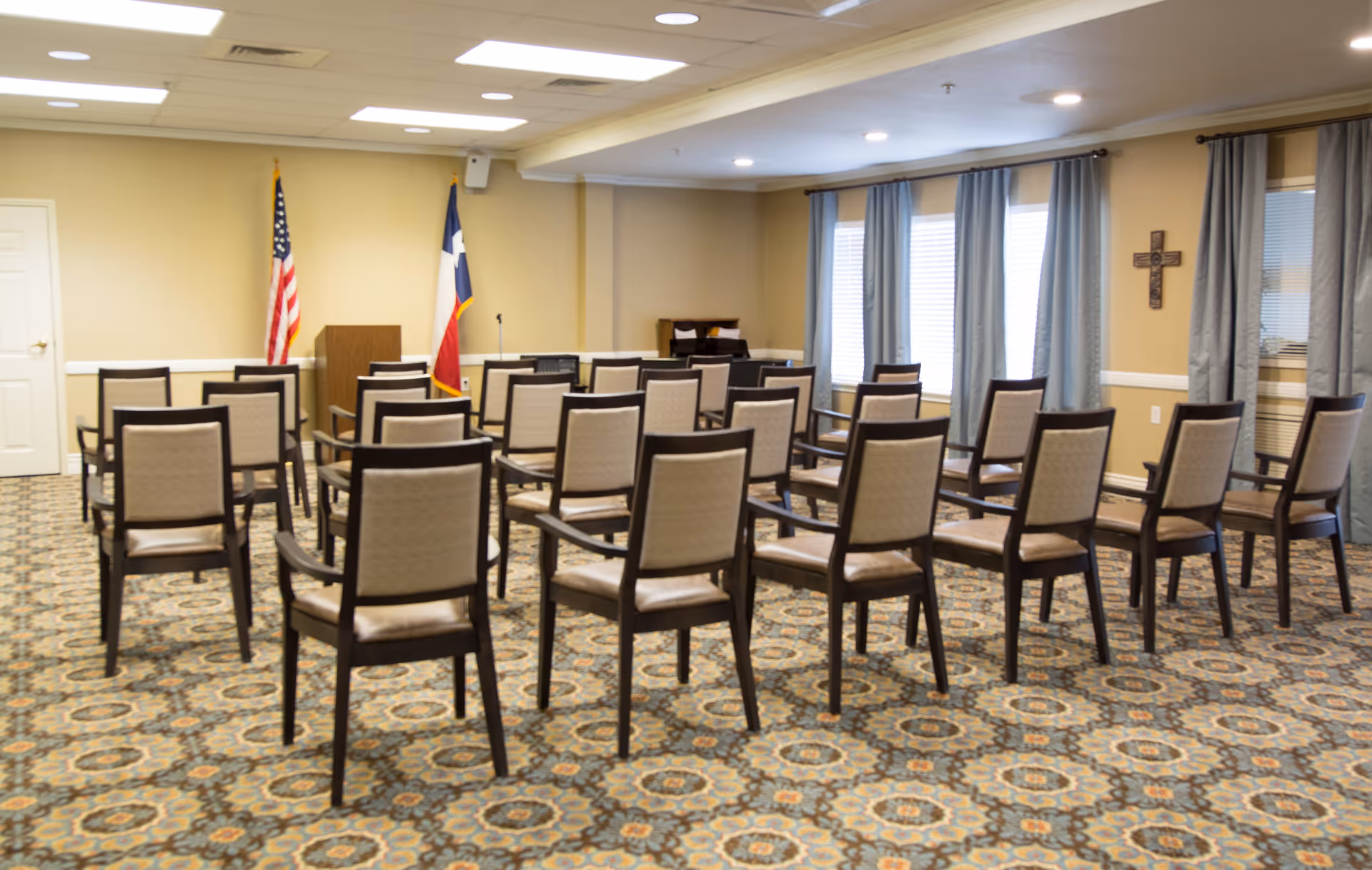 A meeting or conference room with rows of beige cushioned chairs arranged facing a wooden podium. The room has patterned carpet flooring, beige walls, two flags (American and Texas) behind the podium, windows with blue curtains, and a decorative cross on the wall.