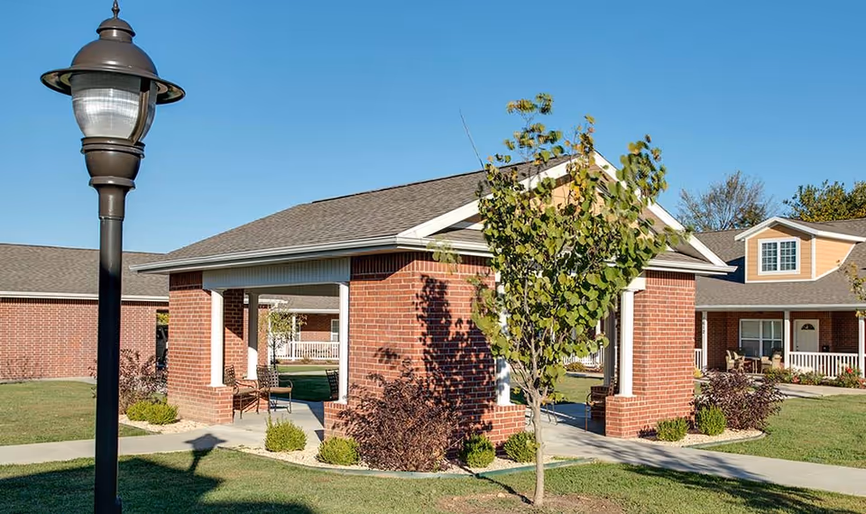 Outdoor view of a senior living facility courtyard with a brick pavilion structure, a black lamppost, green grass, small trees, and shrubs under a clear blue sky.