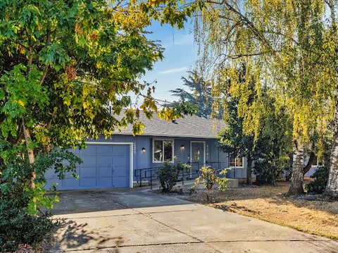 A single-story residential house with a blue exterior, a two-car garage, and a driveway. The house is surrounded by trees and bushes, with a clear blue sky in the background.