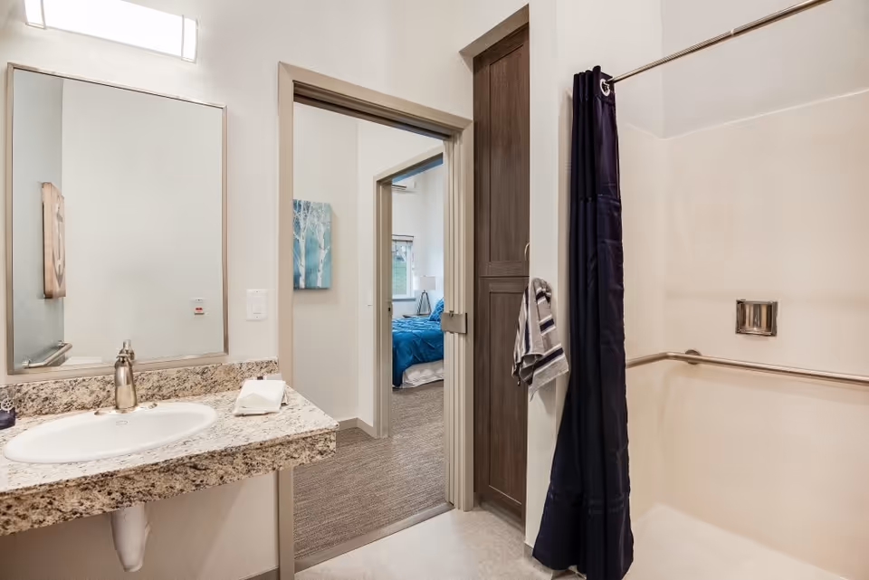 Bathroom with a granite countertop sink, a large mirror, a towel hanging on a hook, and a shower area with a dark shower curtain. The bathroom opens into a bedroom with a blue bedspread and a window.