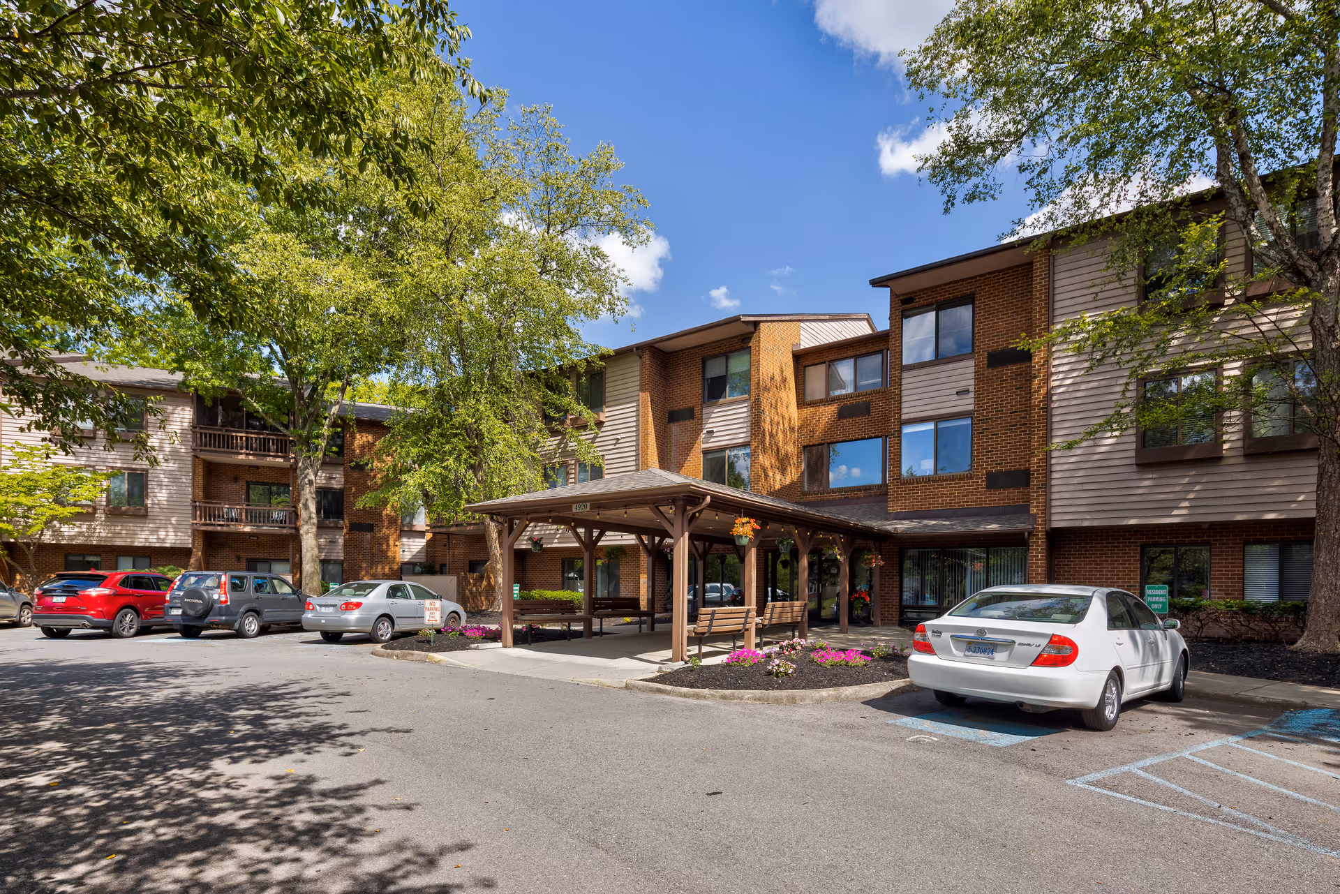 Exterior view of The Park Oak Grove senior living facility showing a three-story building with a mix of brick and siding. There is a covered entrance with benches and hanging flower baskets. Several cars are parked in the parking lot, and trees with green leaves surround the area under a blue sky with some clouds.