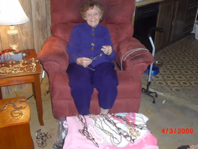 An elderly woman sitting in a red armchair in a room with wood-paneled walls. She is wearing a blue outfit and holding a necklace. In front of her is a table covered with a pink cloth displaying multiple necklaces. To her left is a wooden side table with a lamp and more jewelry. The floor has a patterned linoleum section and a concrete section. A walker and a blue pet bowl are visible in the background.