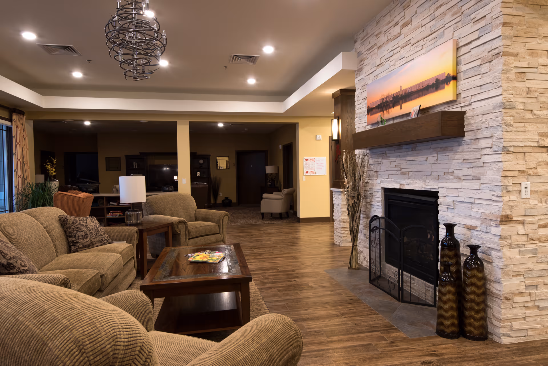 A cozy living room area with beige upholstered sofas and armchairs arranged around a wooden coffee table. The room features a stone fireplace with a wooden mantel, decorated with two tall vases and a panoramic sunset photograph above it. The flooring is wood, and the ceiling has recessed lighting and a decorative chandelier. In the background, there is additional seating and a TV area.