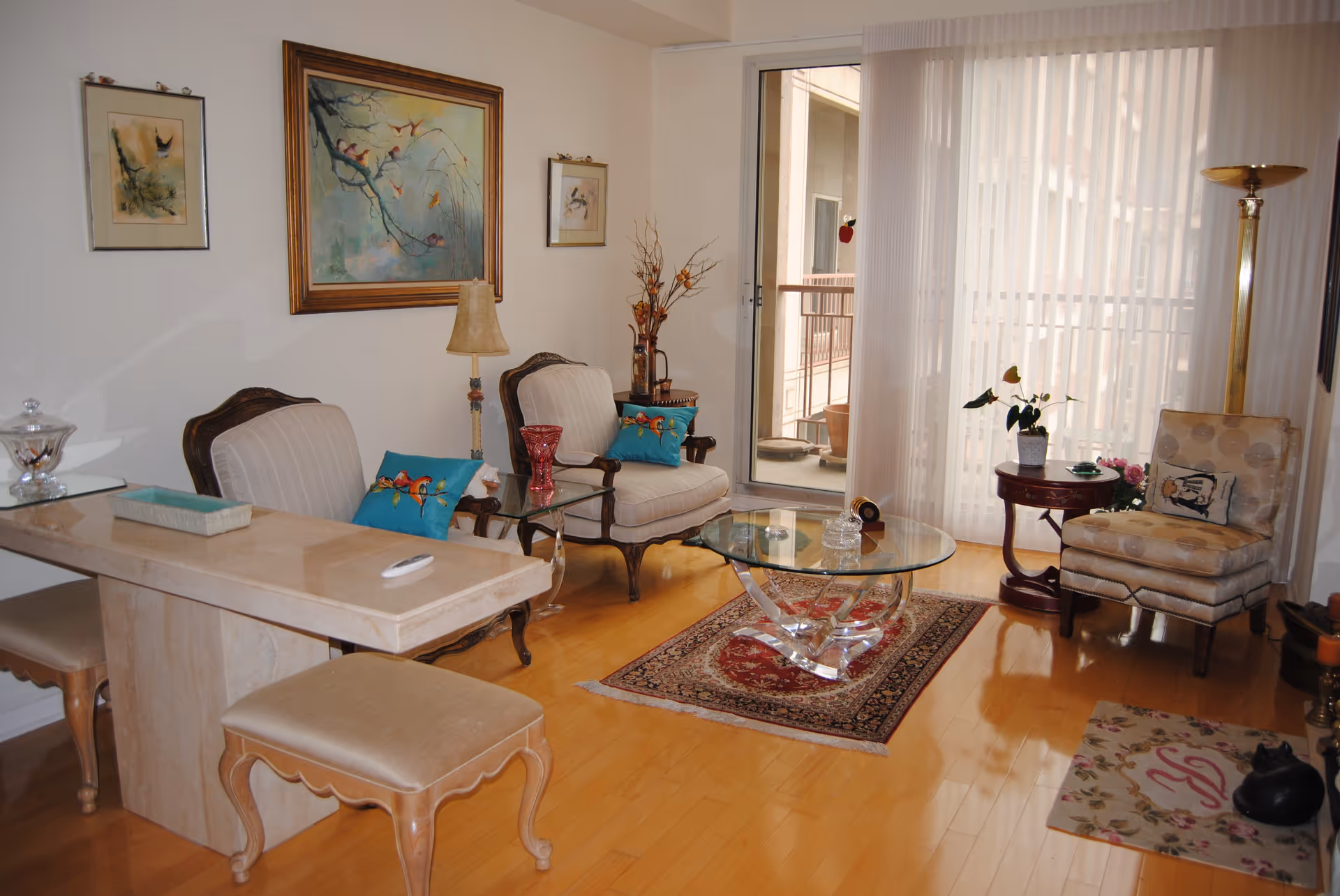 A cozy living room with wooden flooring, featuring two upholstered armchairs with blue bird-themed cushions, a glass coffee table on a patterned rug, a small round side table with a plant, a beige patterned chair, a tall floor lamp, and a marble-topped table with two stools. The room has large sliding glass doors with sheer curtains leading to a balcony. Three framed paintings hang on the walls.