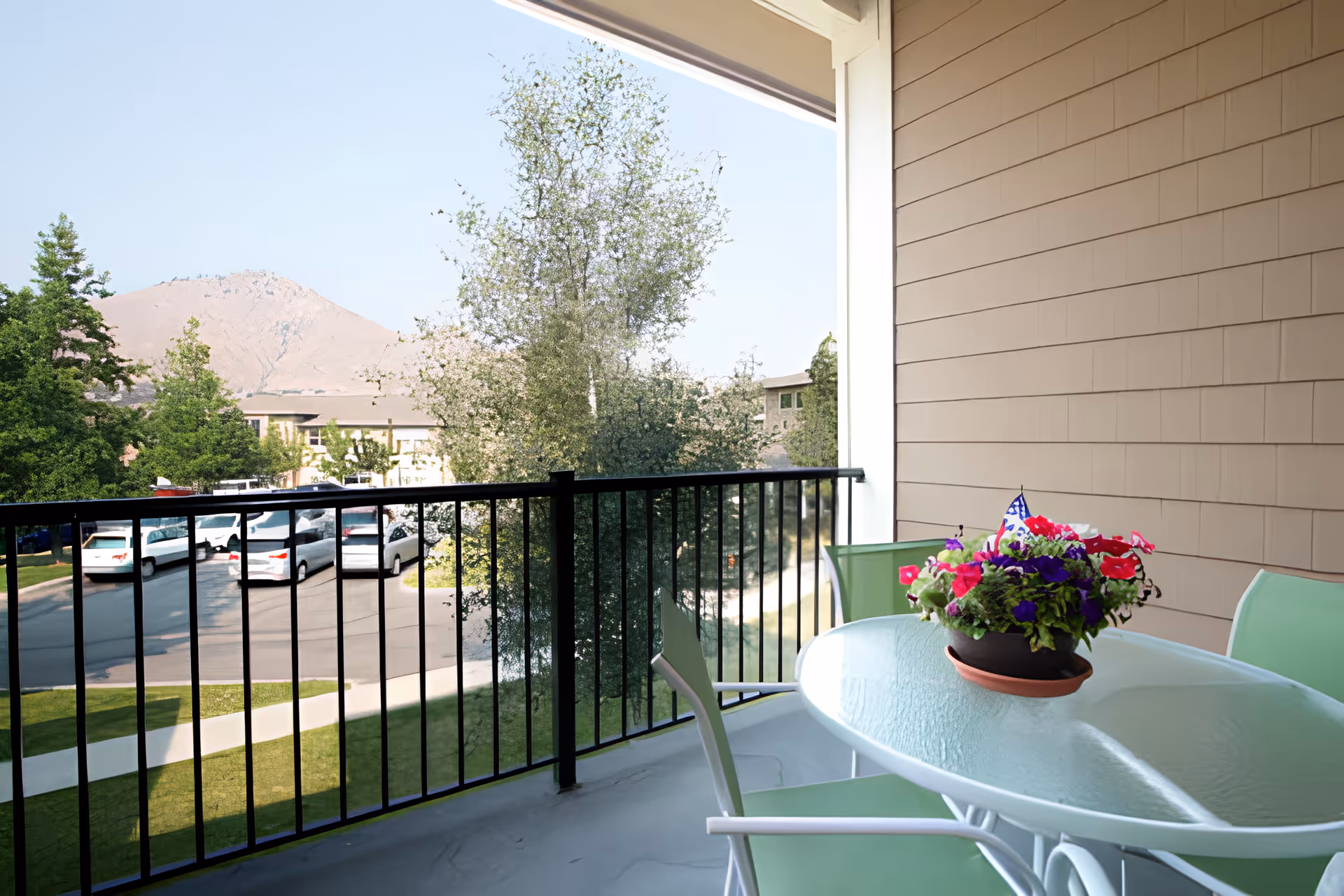 A balcony with a glass-top table and green chairs, featuring a colorful flower pot on the table. The balcony has a black metal railing and overlooks a parking area with several white vehicles, trees, and a mountain in the background under a clear sky.