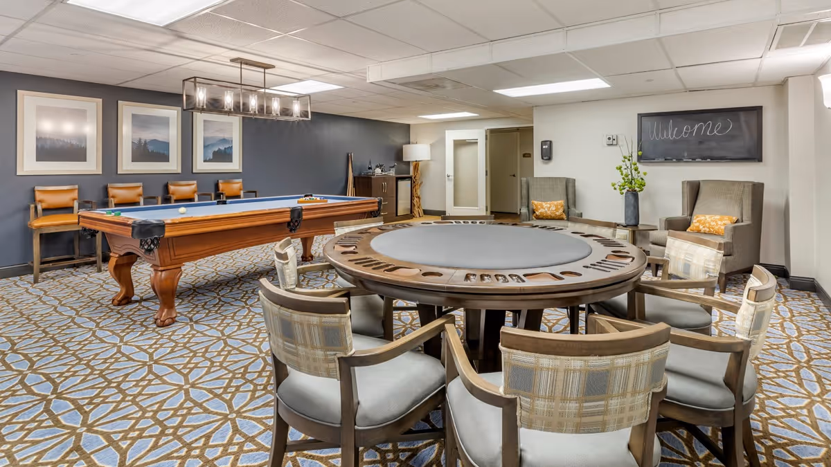 A recreational room with a round game table surrounded by six cushioned chairs, a pool table with balls and cues, three framed landscape pictures on the wall, and two armchairs with yellow pillows near a small table with a vase of flowers. A chalkboard on the wall says 'Welcome'.