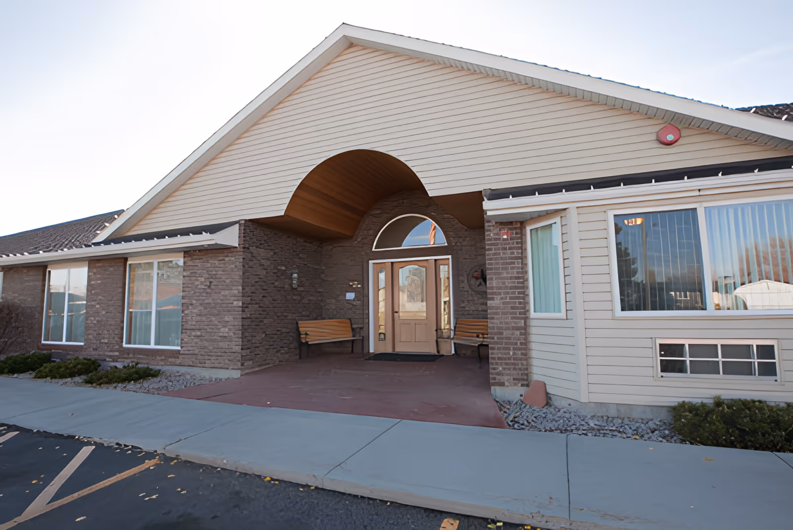 Exterior front entrance of a single-story assisted living facility with a covered porch area featuring two wooden benches, large windows, and a glass door under a peaked roof.