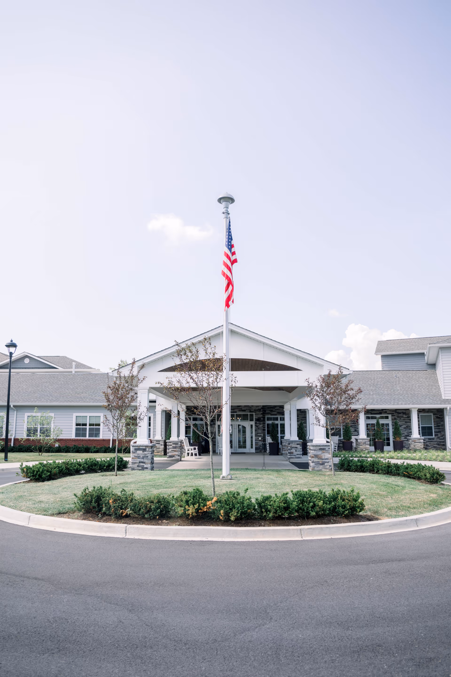 Front exterior view of a senior living facility with a covered entrance, an American flag on a flagpole in the center, small trees, and neatly trimmed bushes surrounding the driveway.