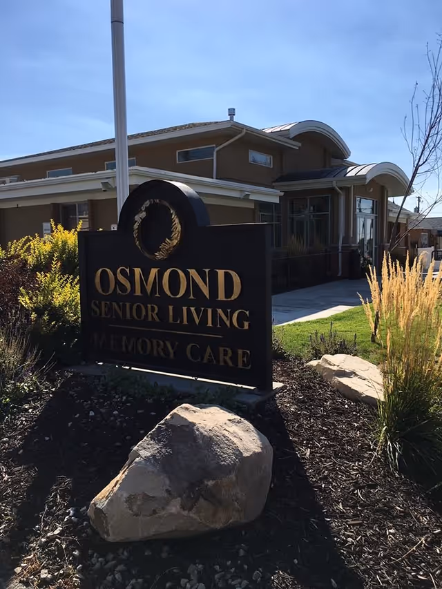 Outdoor view of a senior living facility with a sign that reads 'Osmond Senior Living Memory Care' in front of a modern building with landscaping including rocks, bushes, and ornamental grasses.