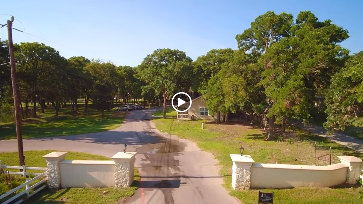 Gated driveway flanked by stone pillars leading to a tree-lined property with a single-story building in the background.
