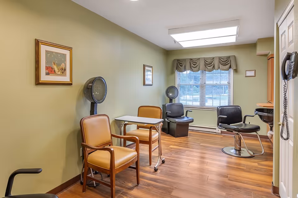 A small salon room with two vintage hair dryers, two tan chairs with a small white table between them, a black salon chair, a black sink, a wall-mounted phone, and a window with green valance curtains. The walls are painted light green and the floor is wood.