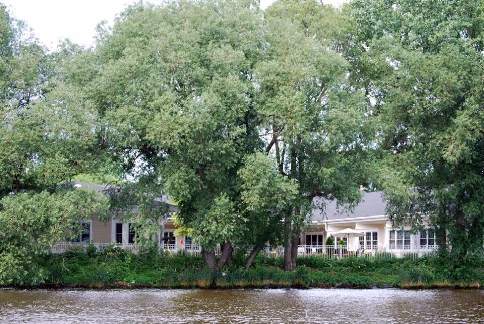 View of a single-story building partially obscured by large leafy trees, situated beside a body of water with greenery along the shoreline.