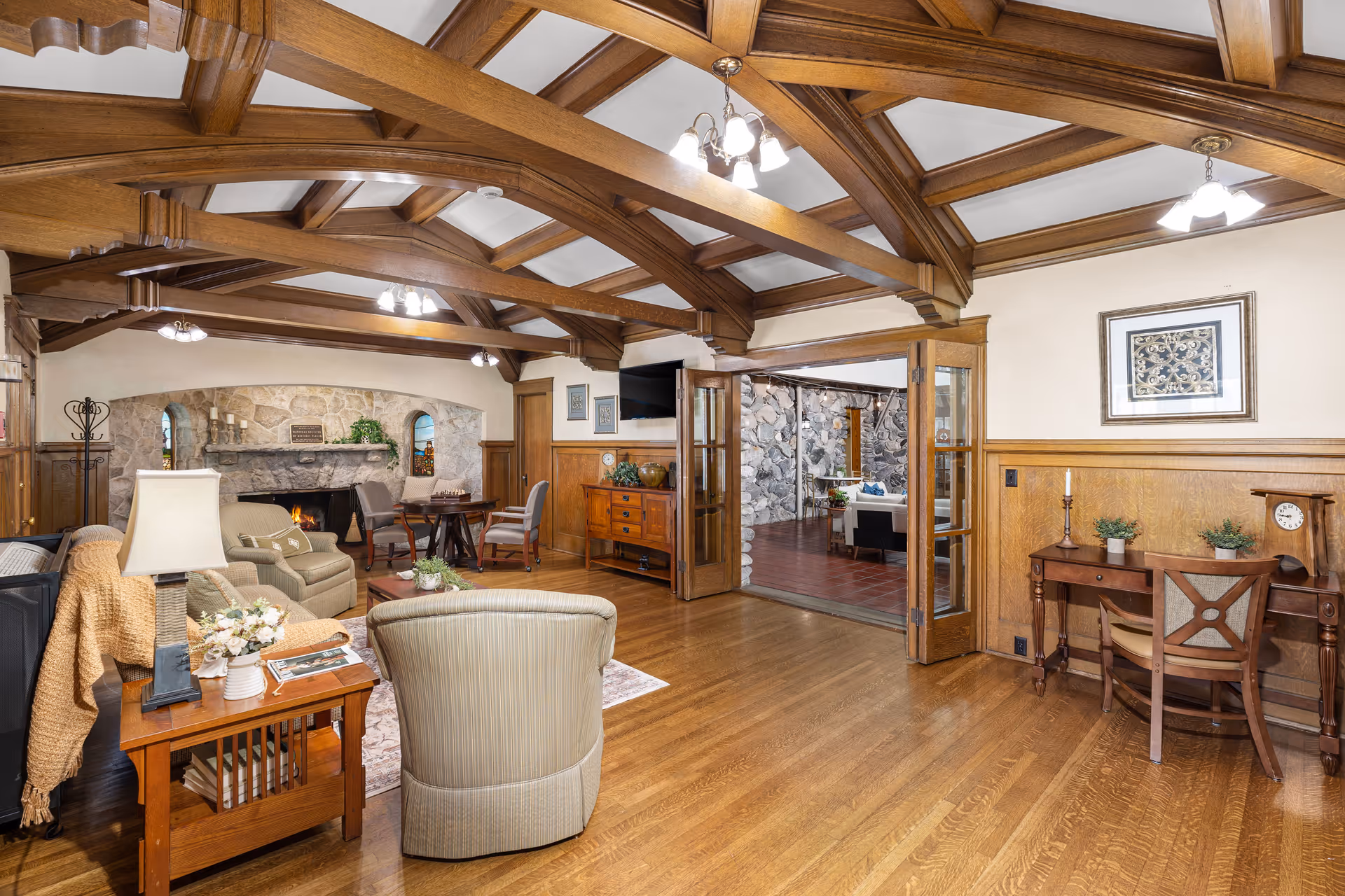 A spacious living room with wooden beams on the ceiling, hardwood floors, and a stone fireplace. The room is furnished with comfortable armchairs, a side table with a lamp and flowers, a wooden desk with a chair, and a wooden cabinet with a TV mounted above it. French doors open to another room with stone walls and additional seating.