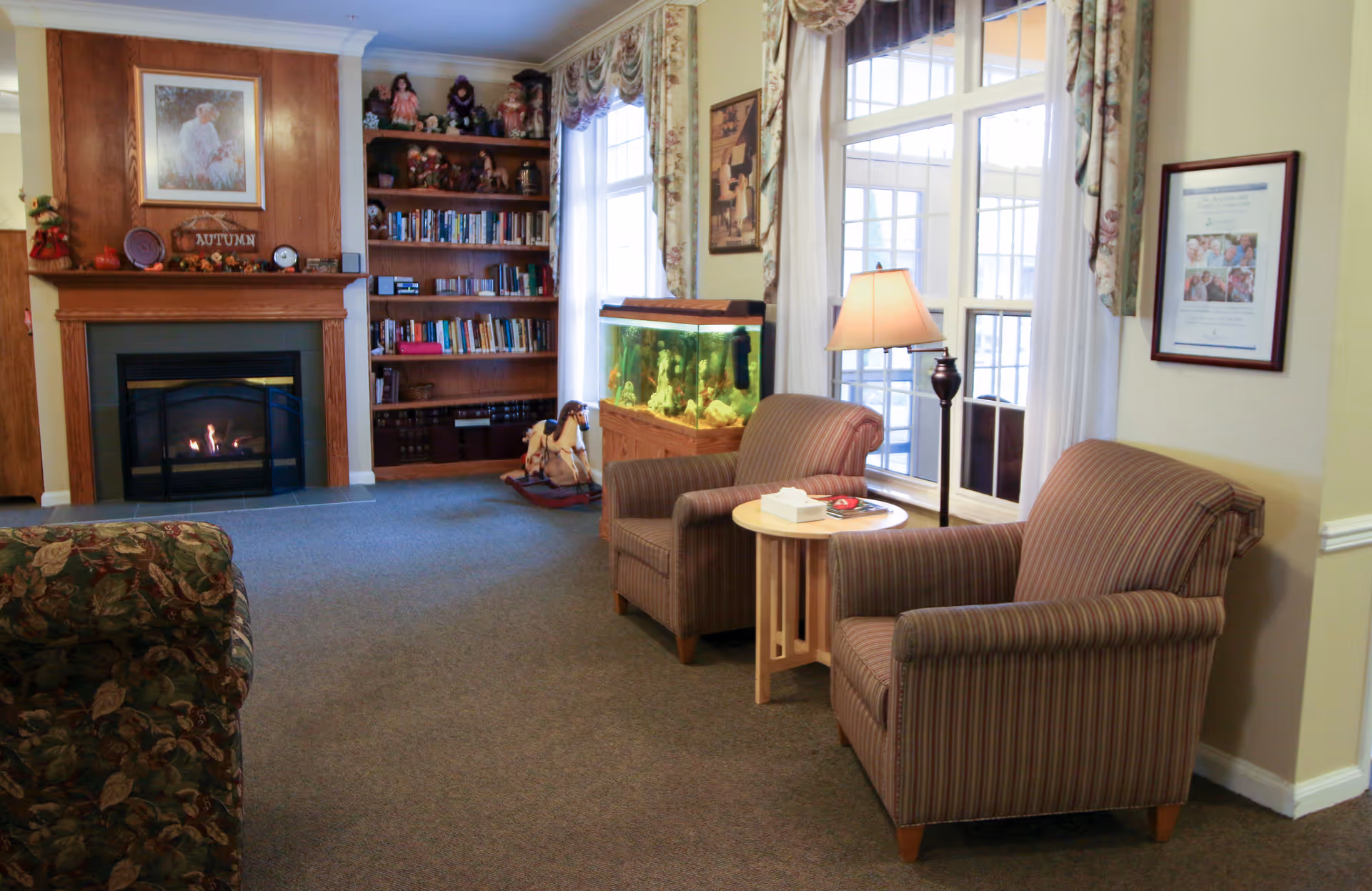 A cozy living room area with two striped armchairs and a small round wooden table between them holding a tissue box and a magazine. There is a floor lamp beside the chairs and a large fish tank near the window with floral curtains. A wooden bookshelf filled with books and decorative dolls is next to a fireplace with a wooden mantel decorated with autumn-themed items and a framed picture above it. The room has carpeted flooring and light-colored walls.