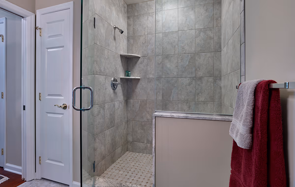 Tiled walk-in shower with a glass door, built-in corner shelves, a half wall, and towels hanging on a towel rack nearby.