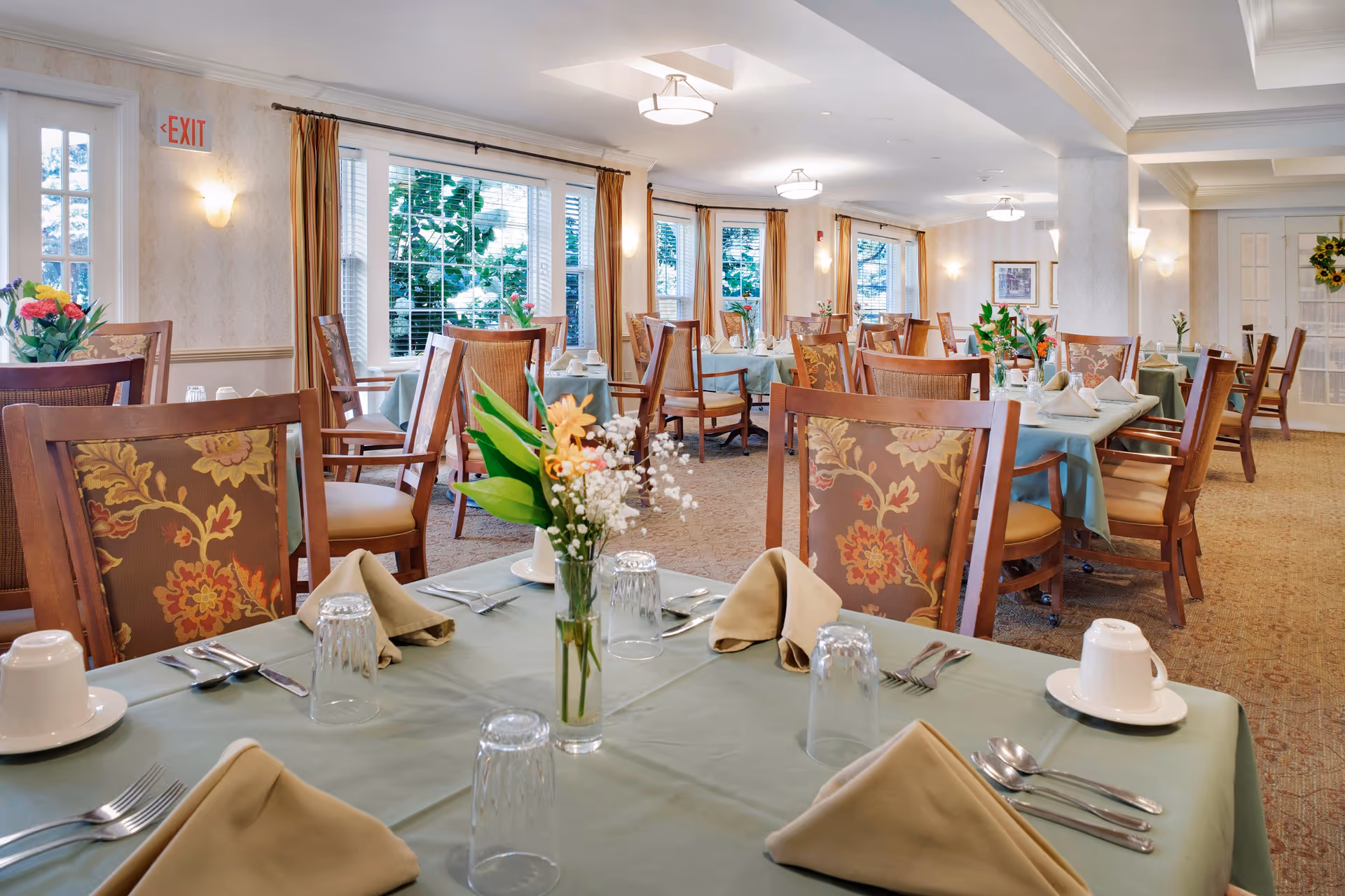 A bright and elegant dining room in a senior living facility with multiple tables covered in light green tablecloths, each set with beige napkins, glassware, silverware, and floral centerpieces. The room features large windows with beige curtains, patterned carpet, and warm lighting fixtures on the ceiling and walls.