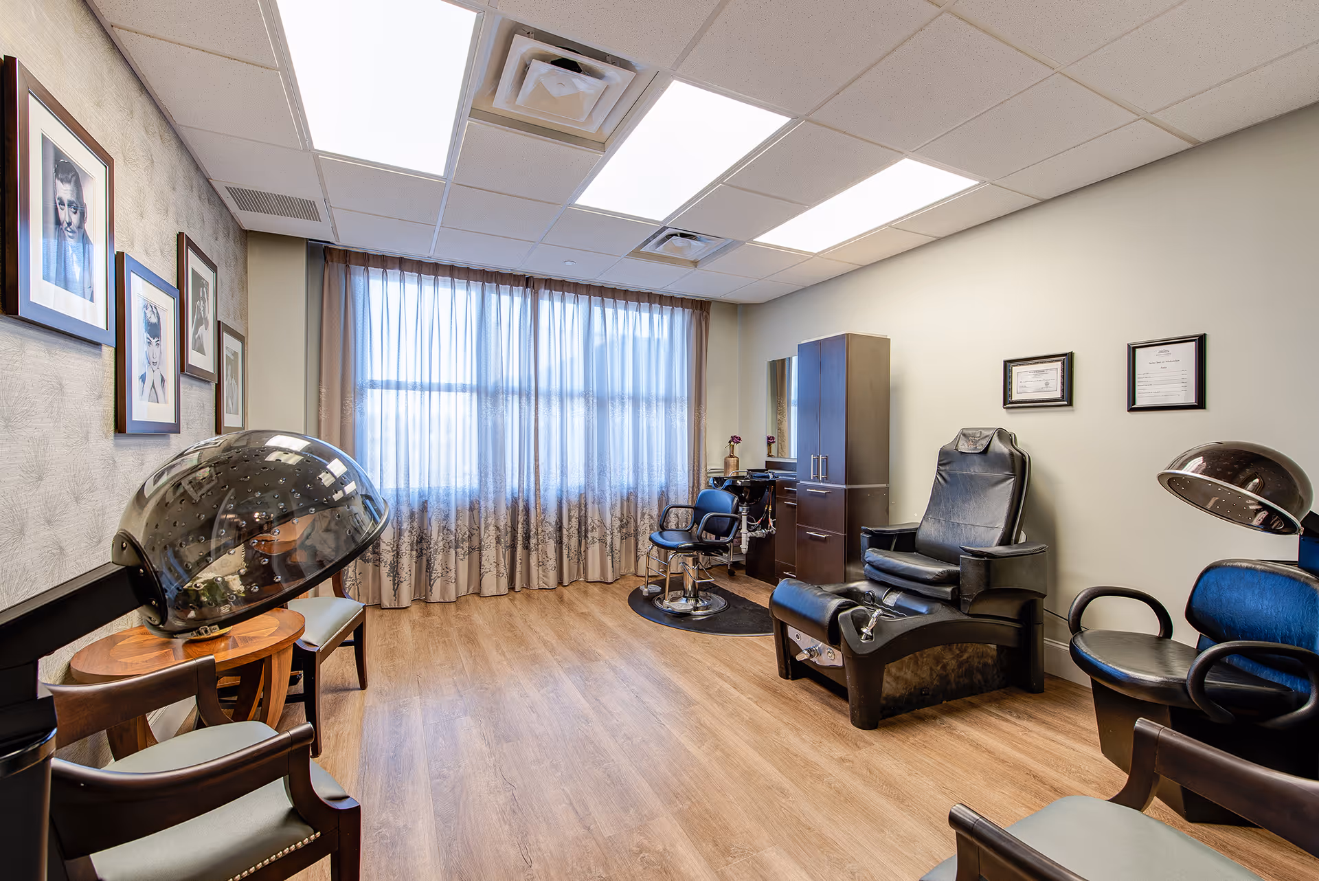 A well-lit salon room with wooden flooring and large windows covered by sheer curtains. The room contains salon chairs, hair drying stations, a pedicure chair, a cabinet with a mirror, and framed black and white portraits on the wall.