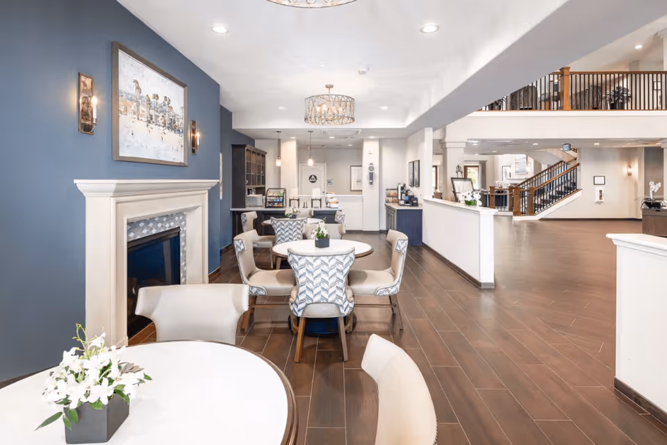 A bright and spacious senior living common area with round tables and cushioned chairs arranged for socializing or dining. The room features a fireplace with a decorative mantle on the left wall, blue accent wall with artwork, and modern light fixtures hanging from the ceiling. In the background, there is a kitchenette area with coffee machines and snacks, and a staircase leading to an upper floor.