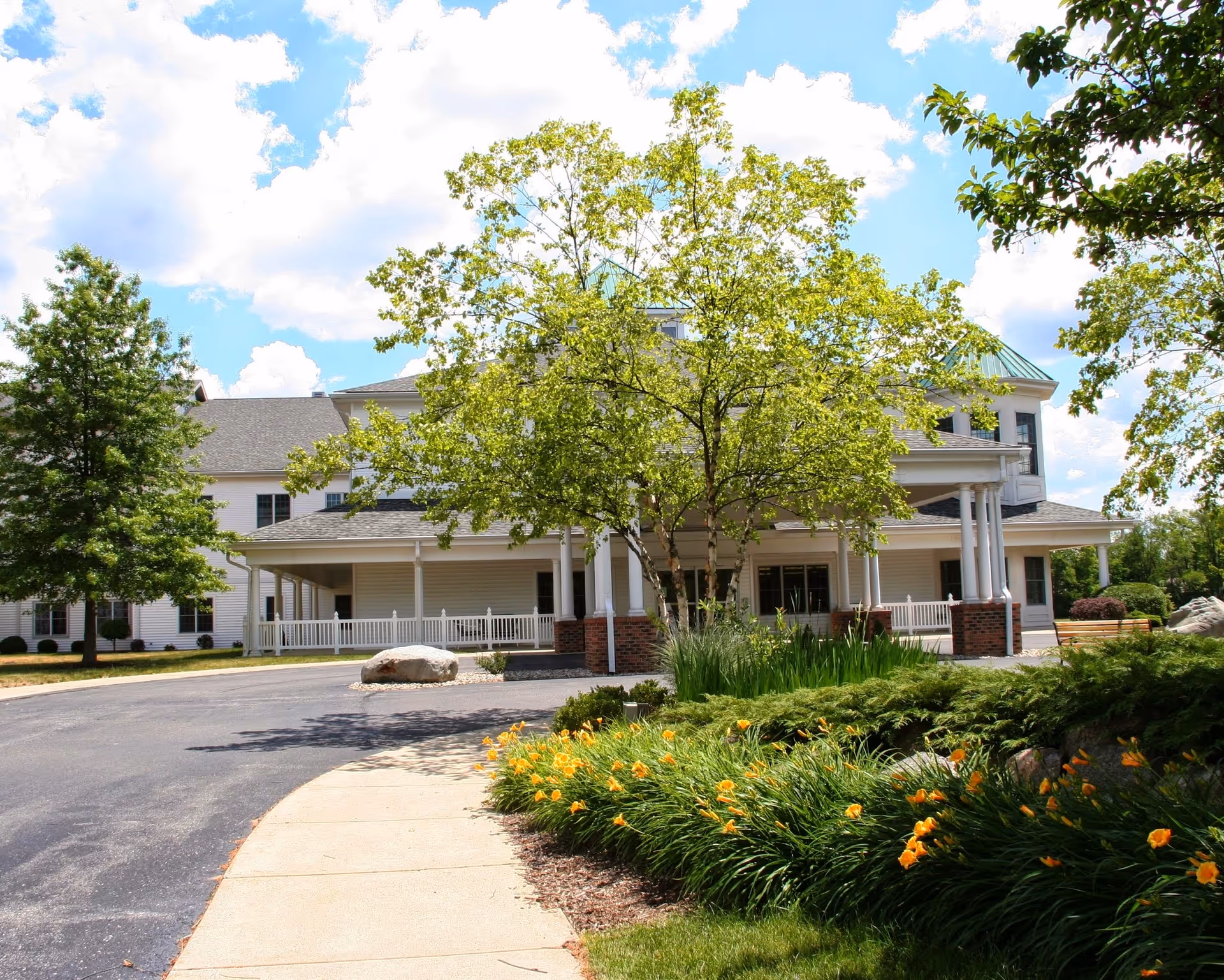 Front entrance of a senior living facility with a circular driveway, porch columns, trees, and landscaped flower beds.