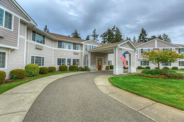 Exterior view of a two-story assisted living facility with beige siding, multiple windows, and a covered entrance with an American flag. The driveway curves in front of the entrance, surrounded by well-maintained green lawns and shrubs under a cloudy sky.